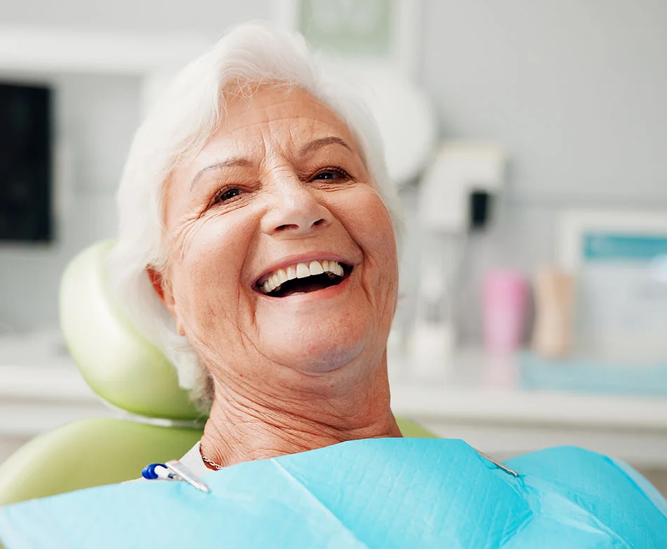 Elderly woman with white hair smiles brightly while sitting in a dentist chair, wearing a blue bib.