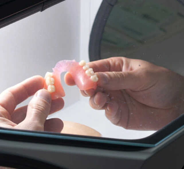 Hands holding a set of pink dental prosthetics with white teeth, viewed through a glass surface, suggesting a clinical setting.
