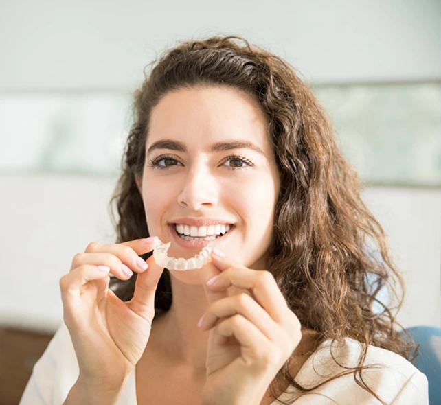 Smiling woman with curly hair holds a clear dental aligner near her mouth, sitting in a bright room.