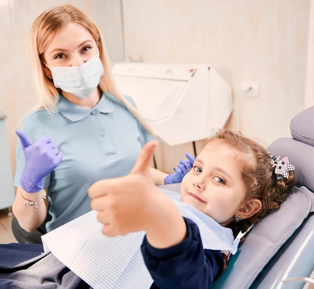 A dentist wearing gloves and a mask sits next to a young girl in a dental chair; both are giving a thumbs up to the camera.