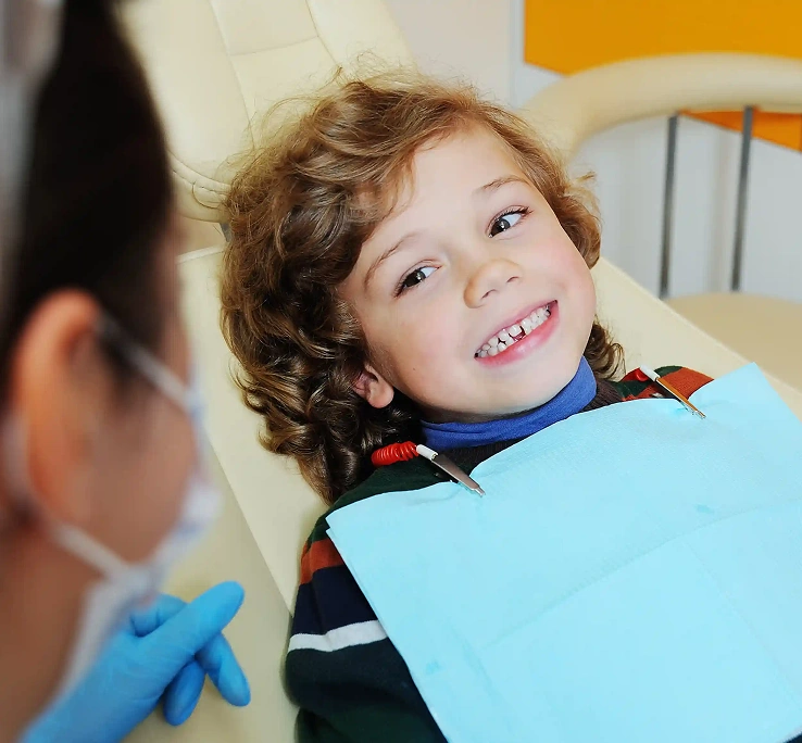 A smiling child with curly hair sits in a dental chair, wearing a blue bib. A dental professional with blue gloves is nearby, creating a cheerful atmosphere.