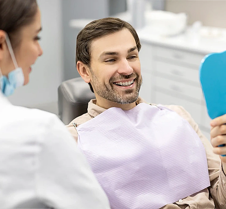 Dentist Narre Warren - A man sits in a dental chair, smiling as he holds a mirror. A dentist, wearing a mask, smiles nearby.