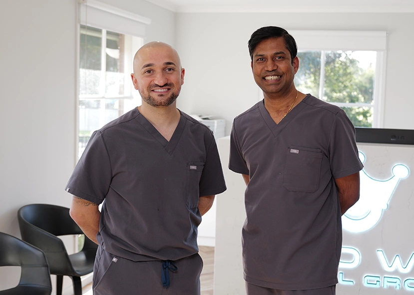Crown Dental Group - Two smiling men in gray scrubs stand in a bright dental office. They exude professionalism and warmth, with a dental group logo partially visible.